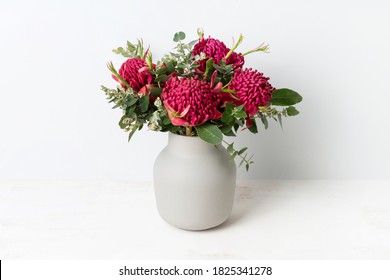 A Bunch Of Australian Native Red Waratah Flowers And Eucalyptus Leaves, In A Stylish Grey Vase On A Table, With A White Background.