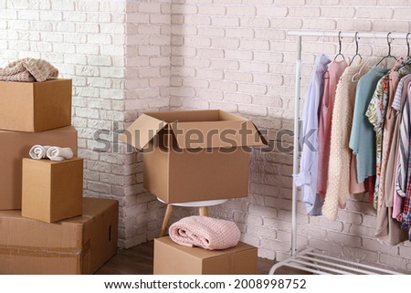 Bunch of blank unsigned moving boxes and clothing hanging on a rack in new empty apartment. Spacious unfurnished room with unpacked cardboard box stack. Interior background, copy space for text.