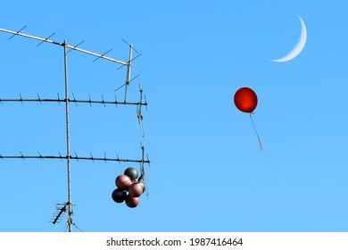 Bunch of colored balloons caught on the roof antenna of the old house. Only single red helium balloon flies up into the blue sky towards the young moon 