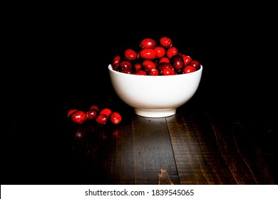 A Bunch Of Cranberrries In A White Porcelain Bowl, With Black Background And Brown Wood Base. Shot In Low Key Style.