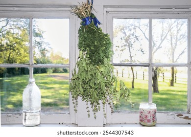 A bunch of drying herbs hangs in front of a vintage white-framed window, flanked by a glass bottle and a floral candle, overlooking a serene countryside scene. - Powered by Shutterstock