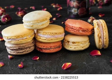 Bunch of french macarons in different colors. Front view, black background. Rose petals and small flower buds as decoration. Selective focus, natural light
