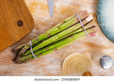 Bunch of fresh green asparagus tied with rubber band placed on wooden table with knife and olive oil, ready for cooking - Powered by Shutterstock