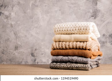 Bunch Of Knitted Warm Pastel Color Sweaters With Different Knitting Patterns Folded In Stack On Brown Wooden Table, Grunged Texture Wall Background. Fall Winter Season Knitwear. Close Up, Copy Space