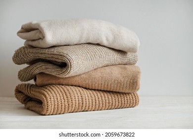 Bunch of knitted warm pastel color sweaters with different knitting patterns stacked in messy pile on white wooden table, white wall background. Fall winter season knitwear. Close up, copy space.