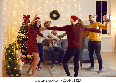 Bunch Of Mixed Race Millennial Friends Reunite At Festive Get Together At Home. Group Of Happy Multiracial Multiethnic People At Christmas Party Meeting Friend And Spreading Arms Wide Open For A Hug