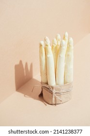 a bunch of raw white asparagus placed in upright standing, vertical position, tied with paper and rope on a two-tone brown-colored background. Hard light projecting shadows
