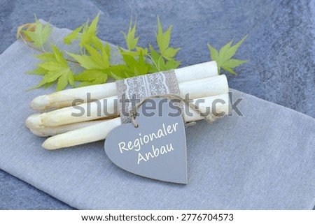 A bundle of white asparagus spears wrapped in lace and jute ribbon, with a gray heart-shaped tag bearing the inscription “Regionaler Anbau.” 
 The German translation is Regional cultivation.
