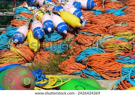 Bundles of commercial nylon woven fishing line orange, yellow, blue, and green color. There are multiple pink buoys piled on top of the rope. The fishing gear is in a pile on a concrete platform. 