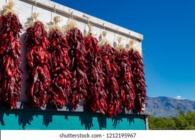 Bundles Of Fresh Ripe Red Hatch Chili Peppers Freshly Picked And Hanging To Dry On The Side Of A White Board With A Turquoise Panel, With Mountains And A Blue Sky Background. Roadside Vendor Display. 