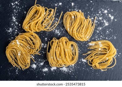 Bundles of uncooked spaghetti are artistically arranged on a textured black slate surface, lightly dusted with flour, creating a rustic culinary scene. - Powered by Shutterstock