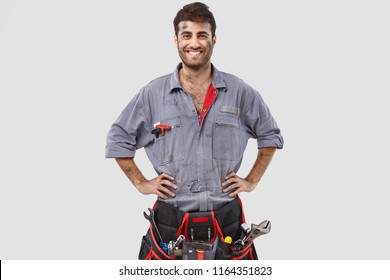 Buoyant Successful Handyman Wears Grey Overalls, Tool Belt, Stands With Hands On Waist Over White Background Has Shining Smile. Content Industrial Worker Rejoices Coming Weekend After Hard Working Day