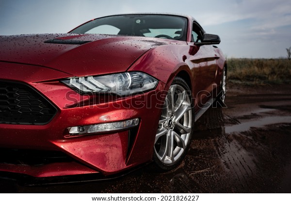 Burgas, Bulgaria - 07\05\2021: Ford Mustang,\
red sport car in a field on a dirt road after rain with drops on\
the body of car, in the photo there are headlights and a side view\
of the car in closeup