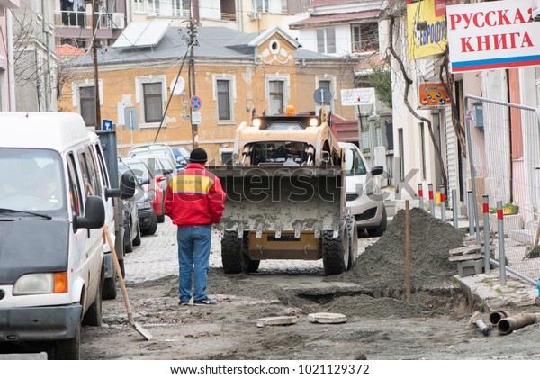 Burgas, Bulgaria - 2.7.18: Construction\
inspector is checking the outgoing work on the reconstruction on\
the street, standing next to a big hole in the ground with\
excavator and houses in\
background.