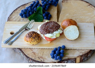 Burger Bun Sandwich With Grilled Meat, Vegetables And Cheese. Home Baked Burger Buns With Flax Seeds On Wooden Board; Grapes And Basket With Vegetables In Background. Bread Texture Of Sliced Bun.