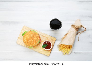 Burger, Chips And Cola. Hamburger And French Fries Wrapped In Brown Wrapping Paper. Fast Food Take Away At White Shabby Chic Wood. Hamburger With Tomato Sauce. Top View, Flat Lay With Copyspace