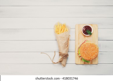 Burger And Chips. Hamburger And French Fries Wrapped In Brown Wrapping Paper. Fast Food Take Away At White Shabby Chic Wood. Hamburger With Tomato Sauce. Top View, Flat Lay With Copyspace