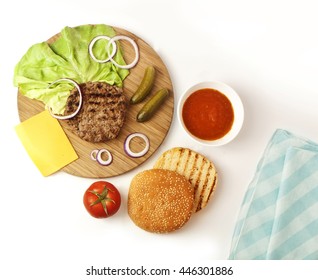 Burger Ingredients: Leaves Of Green Lettuce, Slices Of Cheddar Cheese, Gherkins, Red Onions, Tomatoes, Meat Patty, Grilled Buns With Sesame Seeds, And Tomato Sauce, Shot From Above On White Background