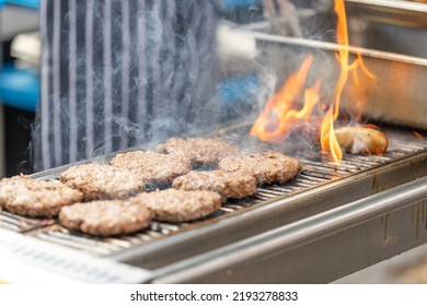 Burger Patties Being Flame Grilled On An Outdoor Bbq. Flames Rising From The Bbq Grill. Chef Apron In The Background Out Of Focus