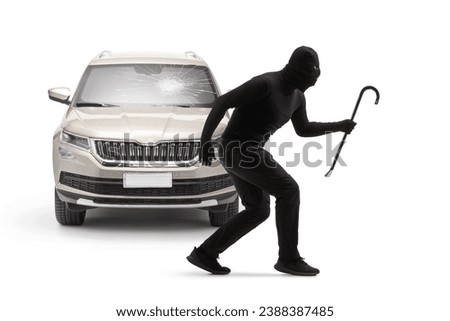 Burglar with balaclava and a crowbar standing in front of a car with a broken windscreen isolated on white background