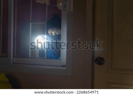 Burglar With Crowbar And Flashlight Looking Into A House Windows.Thief Breaking House Window To Enter House.Burglar with flashlight looking through the window