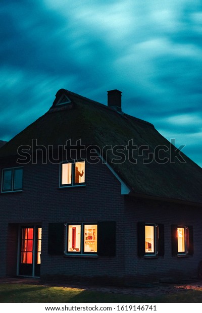 Burglar with handgun in ominous house with
illuminated windows under stormy sky at
dusk.