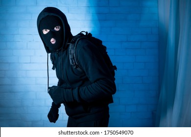 Burglary Concept. Intruder In Black Balaclava Standing In Apartment Or Office In The Dark, Looking At Camera