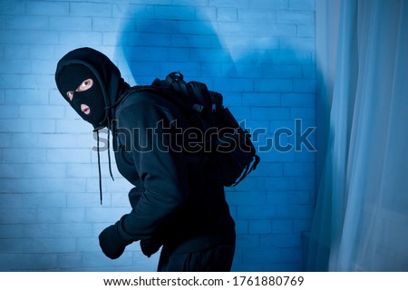 Burglary Concept. Sneaky and scared intruder wearing black balaclava hat lurking in the dark, looking at camera