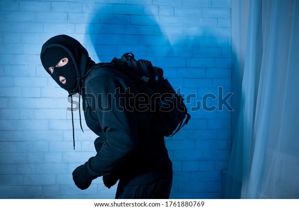 Burglary Concept.\
Sneaky and scared intruder wearing black balaclava hat lurking in\
the dark, looking at\
camera