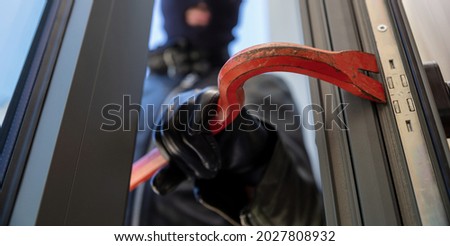 Burglary with crowbar. Burglar in balaclava breaking a glass door. Masked housebreaker entering a house for stealing in the daytime.