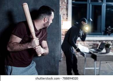 Burglary. Terrified young bearded man hiding and holding a stick while a masked burglar holding a torch and standing at the table and looking for something there