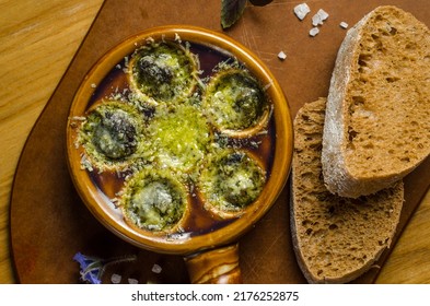Burgundy Snails With Green Butter And Cheese, Gourmet Dish, In Traditional Ceramic Pans With Bread And Glass Of White Wine On A Wooden Table. Dish Close-up. Top View