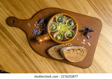 Burgundy Snails With Green Butter And Cheese, Gourmet Dish, In Traditional Ceramic Pans With Bread And Glass Of White Wine On A Wooden Table. Top View