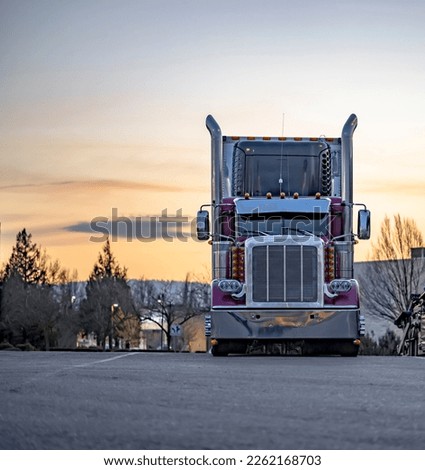 Burgundy stylish big rig American semi truck tractor with high chrome pipes and refrigerator semi trailer standing for the truck driver rest on wide parking lot at twilight evening time with sunset