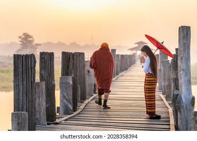 Burmese girls pay homage to the monks passing by on the bridge. Woman wearing traditional clothes with red umbrella at U Bein teak bridge in Mandalay, Myanmar. - Shutterstock ID 2028358394