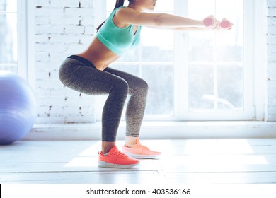 Burn in buttocks. Side view of young woman in sportswear doing squat and holding dumbbells while standing in front of window at gym