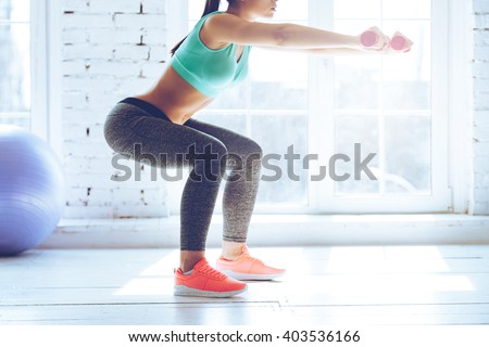 Burn in buttocks. Side view of young woman in sportswear doing squat and holding dumbbells while standing in front of window at gym