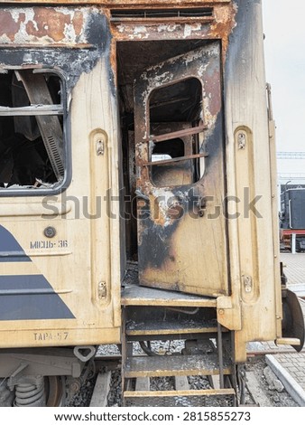 Burned-out train car in rusty condition, deteriorated metal, peeling paint, charred interior, ajar door revealing damage, outdoor railway setting with gravel tracks and other equipment visible, muted