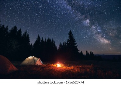 Burning bonfire in tent city without people under bright starry sky with Milky way against the backdrop of silhouettes pine forest and valley of hills. Healthy lifestyle and relaxation concept.