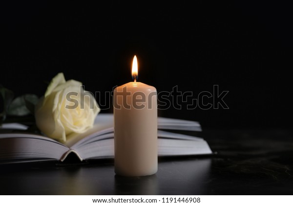 Burning candle, book and white rose on\
table in darkness, space for text. Funeral\
symbol