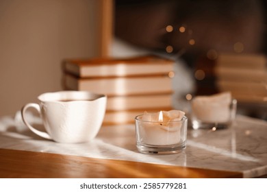 Burning candle with cup of coffee and stack of book on marble table in room. Cozy home atmosphere. Good morning. 
