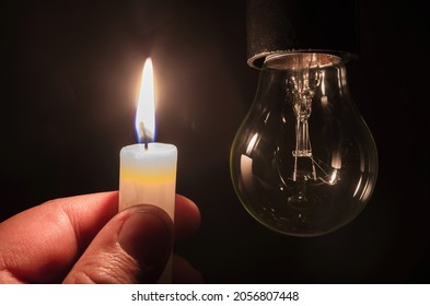Burning candle near a switched off light bulb in complete darkness. Blackout, electricity off, energy crisis or power outage, concept image. 