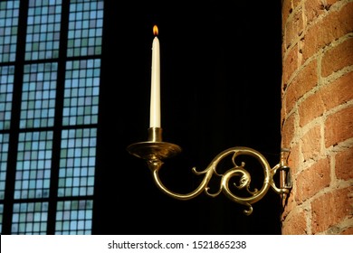 A Burning Candle On A Candle Holder In The Great Church In Deventer, The Netherlands. In The Background The Beautiful Windows With Stained Glass Can Be Seen,