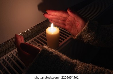 A burning candle with women's hands frozen from frost on the radiator heating in dark home. Shutdown of heating and electricity, power outage, blackout, load shedding or energy crisis, concept image. - Shutterstock ID 2205654561