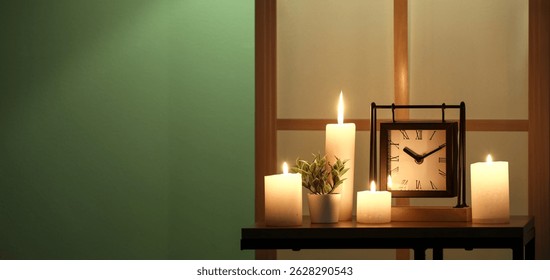 Burning candles and clock on table near green wall in dark room. Banner for design
