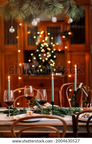 Burning candles and glasses of red wine placed near plates and coniferous branches during Christmas celebration at home