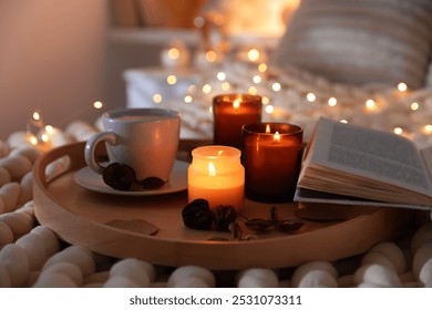 Burning candles, lights, coffee, book and decor on bed indoors, closeup. Autumn atmosphere