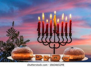 Burning Candles, Menorah And Festive Sweet Donates On Wooden Plates With Dreidels - Hebrew Letters On Four Sides Of Dreidel Means - Great Miracle Happened Here. Blurred Morning Sky And Mountains