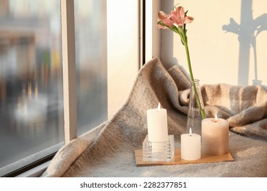 Burning candles and vase with flowers on plaid near window