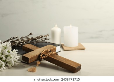 Burning church candles, rosary beads, wooden cross, Bible, willow branches and snowdrops on white table. Space for text